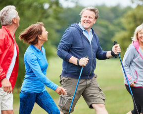 Vier Personen gehen gemeinsam mit Nordic-Walking-Stöcken auf einem Weg in der Natur. Sie tragen sportliche Kleidung und lächeln.