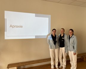 Drei Personen in Arbeitskleidung stehen vor einer Wandprojektion mit dem Wort "Apraxie".