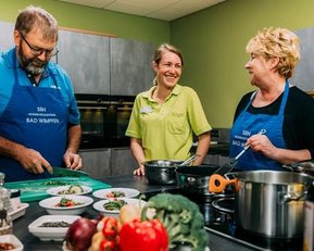Drei Personen in einer Küche bereiten gemeinsam eine Mahlzeit vor. Frisches Gemüse und Obst sind auf dem Tisch angerichtet.