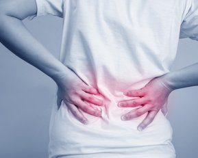 Eine Person hält sich mit beiden Händen den unteren Rücken, der leicht rot hervorgehoben ist, was auf Schmerzen hinweist.