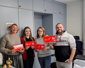 Robert Koch Krankenhaus Stuttgart zu Besuch Vier Personen stehen in einem Büro und halten rote Verpackungen in den Händen. Im Hintergrund sind Büromöbel und ein Kalender sichtbar.