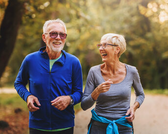 Ein älteres Paar joggt gemeinsam auf einem Weg im Freien, beide lachen und tragen sportliche Kleidung.