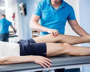 Chronischer Schmerz Ein Physiotherapeut behandelt das Bein eines Patienten auf einer Liege, während ein weiterer Patient im Hintergrund an einer Übung arbeitet.