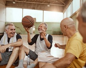 Vier ältere Männer sitzen auf dem Boden eines Basketballfeldes, lachen und unterhalten sich, während einer einen Basketball in der Hand hält.