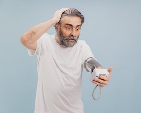 Ein älterer Mann mit grauem Bart und Haar, der ein Blutdruckmessgerät an seinem Arm trägt, schaut überrascht und besorgt auf das Display des Geräts, während er sich mit der anderen Hand den Kopf hält.
