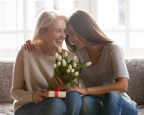 Eine Frau überreicht ihrer Mutter einen Geschenkkarton, während sie gemeinsam mit einem Strauß Tulpen auf einem Sofa sitzt.
