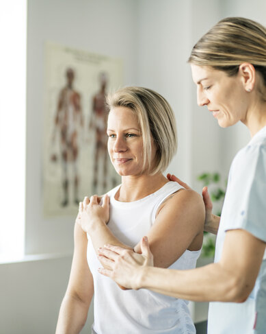 Eine Physiotherapeutin führt eine Behandlung an einer Patientin durch, die lächelnd auf einem Stuhl sitzt. Im Hintergrund sind anatomische Modelle sichtbar.